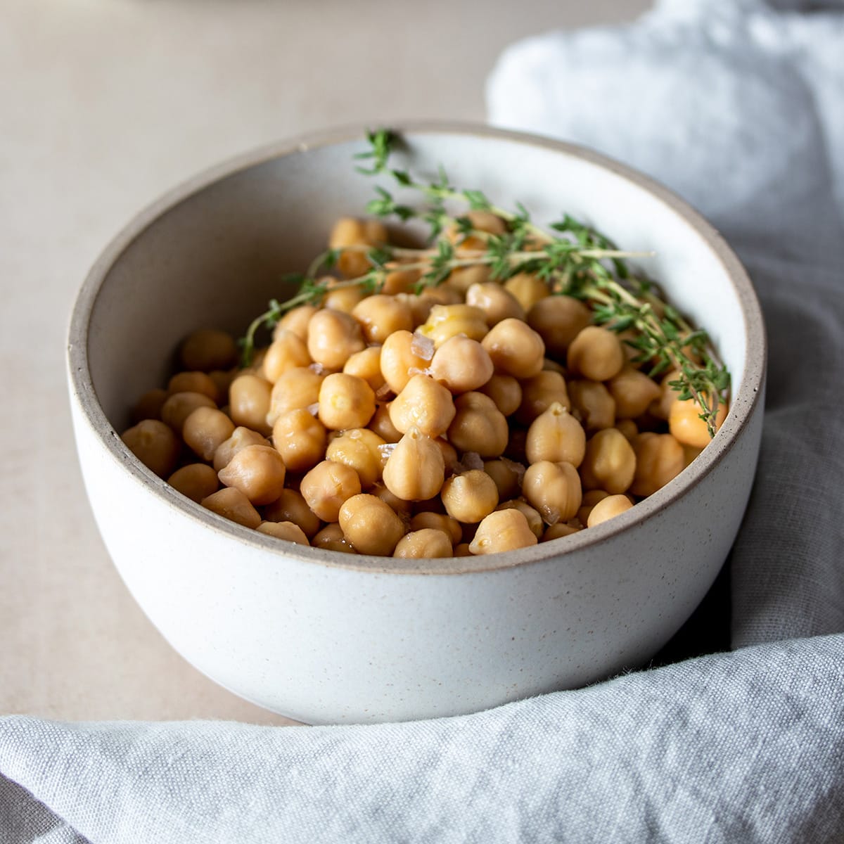 Cooked chickpeas in a white bowl with tan rim and sprigs of thyme and a grey towel around it.