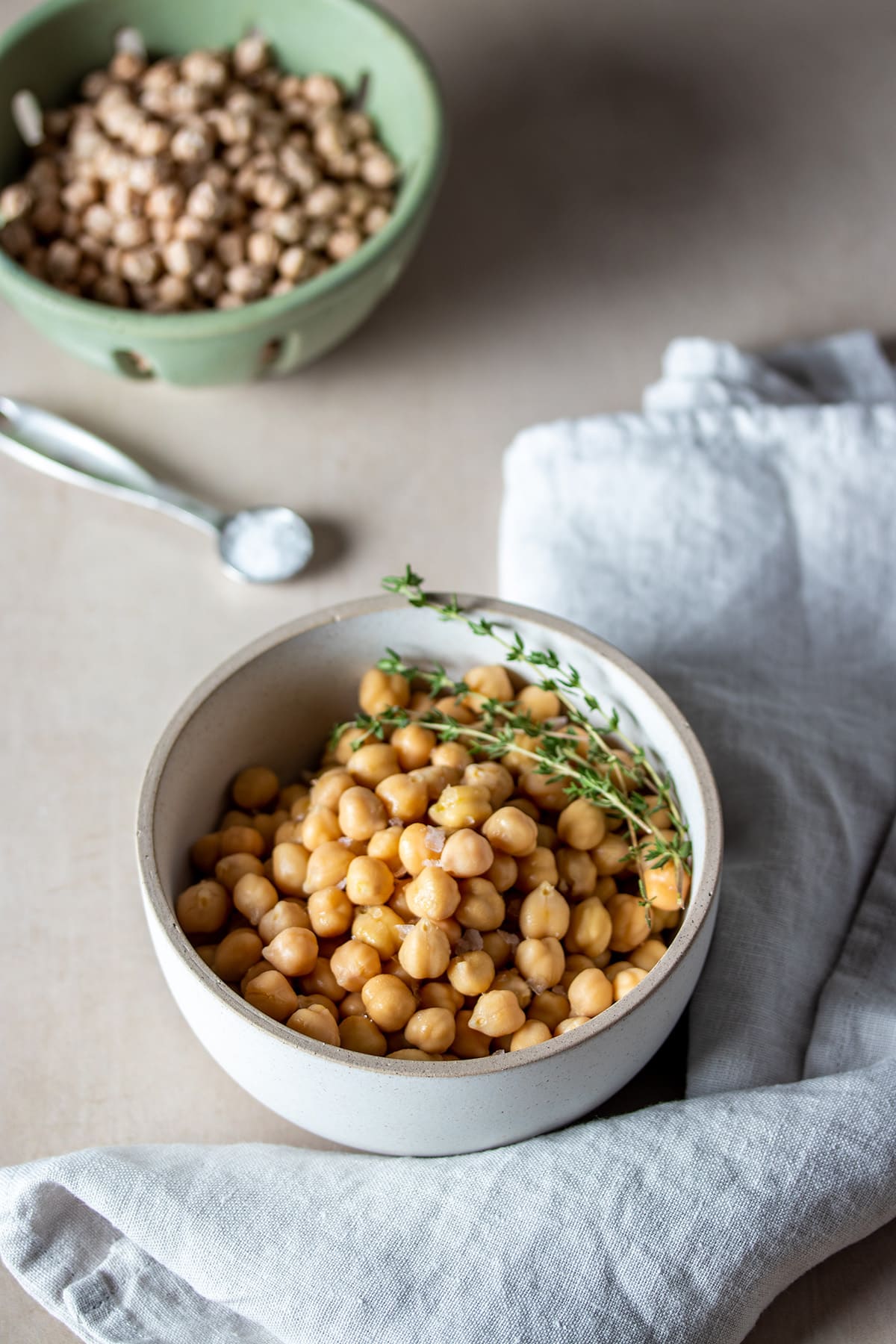 A white bowl filled with chickpeas and thyme sprigs on a tan surface in front of a green strainer with dry chickpeas.