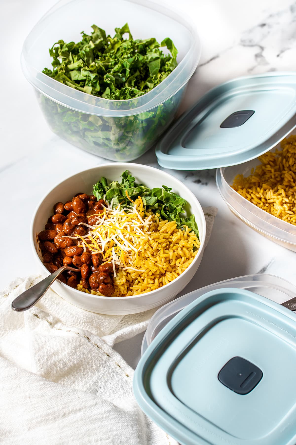 Plastic containers with blue lids and greens and rice in two next to a bowl with beans, rice and greens in it.