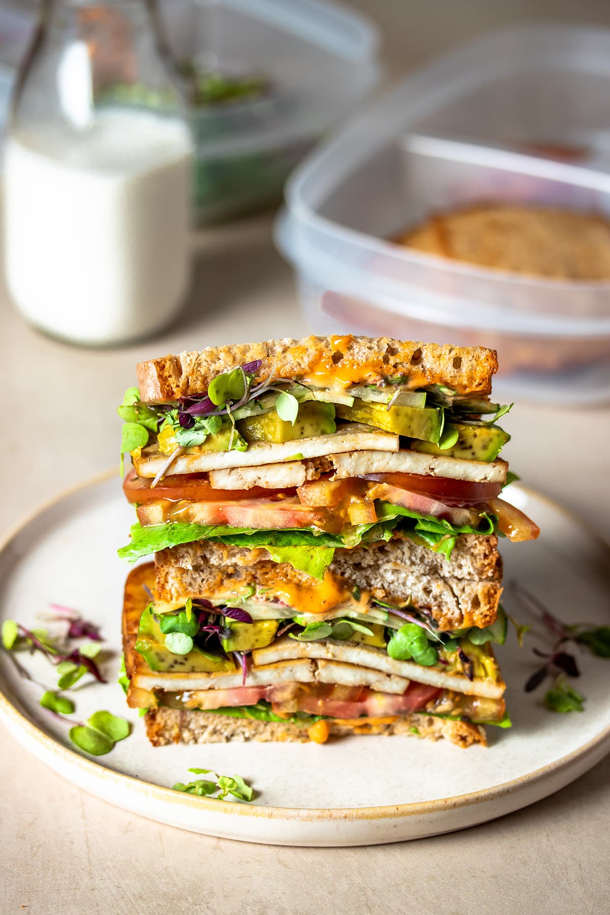 A stacked tofu and veggie sandwich that has been cut in half sitting on a plate with a glass of milk and plastic containers behind it.