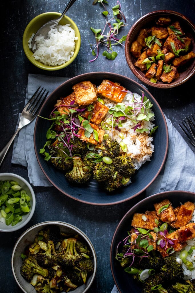 A bowl with baked teriyaki tofu and broccoli over rice with bowls of extra ingredients around it.
