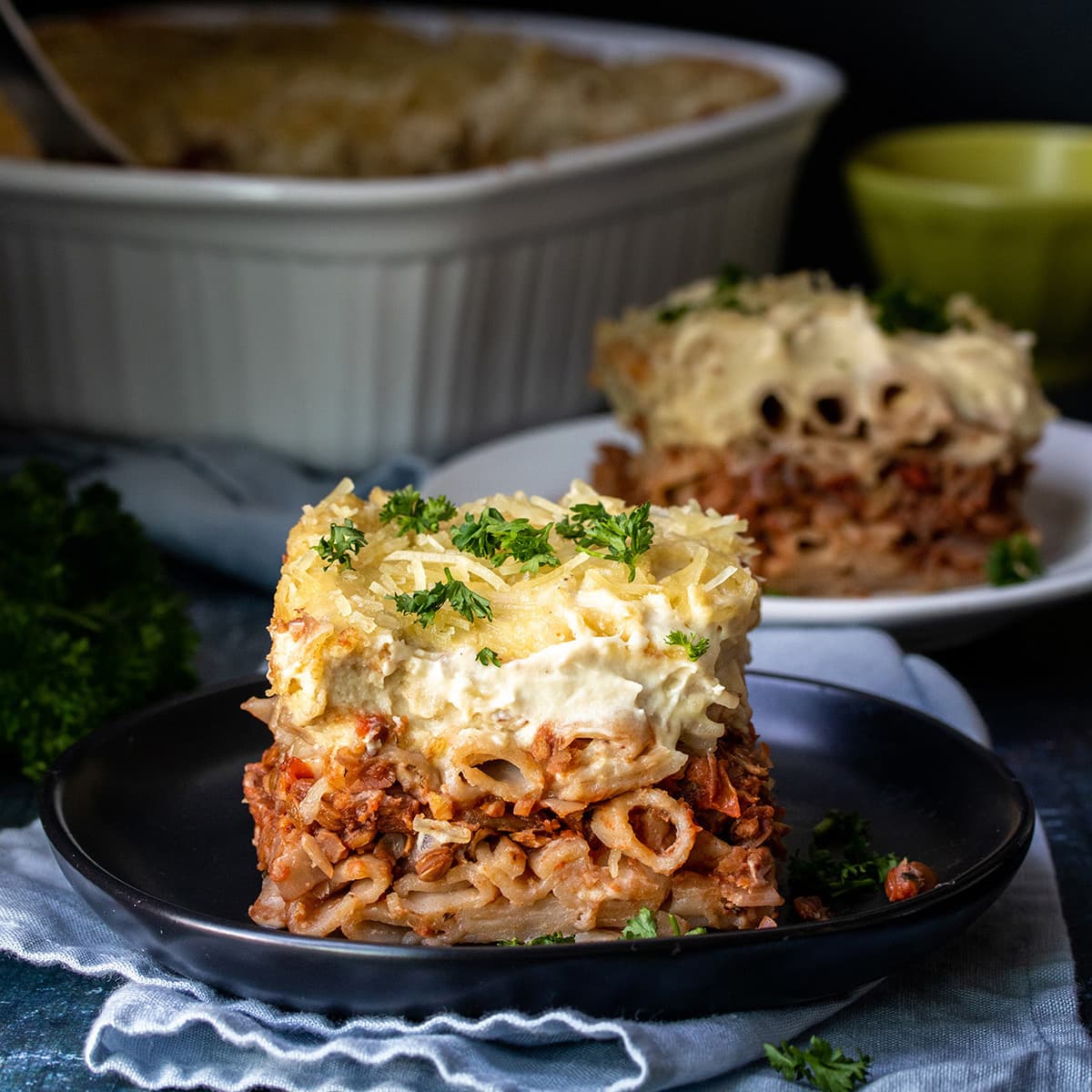 Front view of a piece of pastitsio on a black plate sprinkled with parsley and more pastitsio in the background.