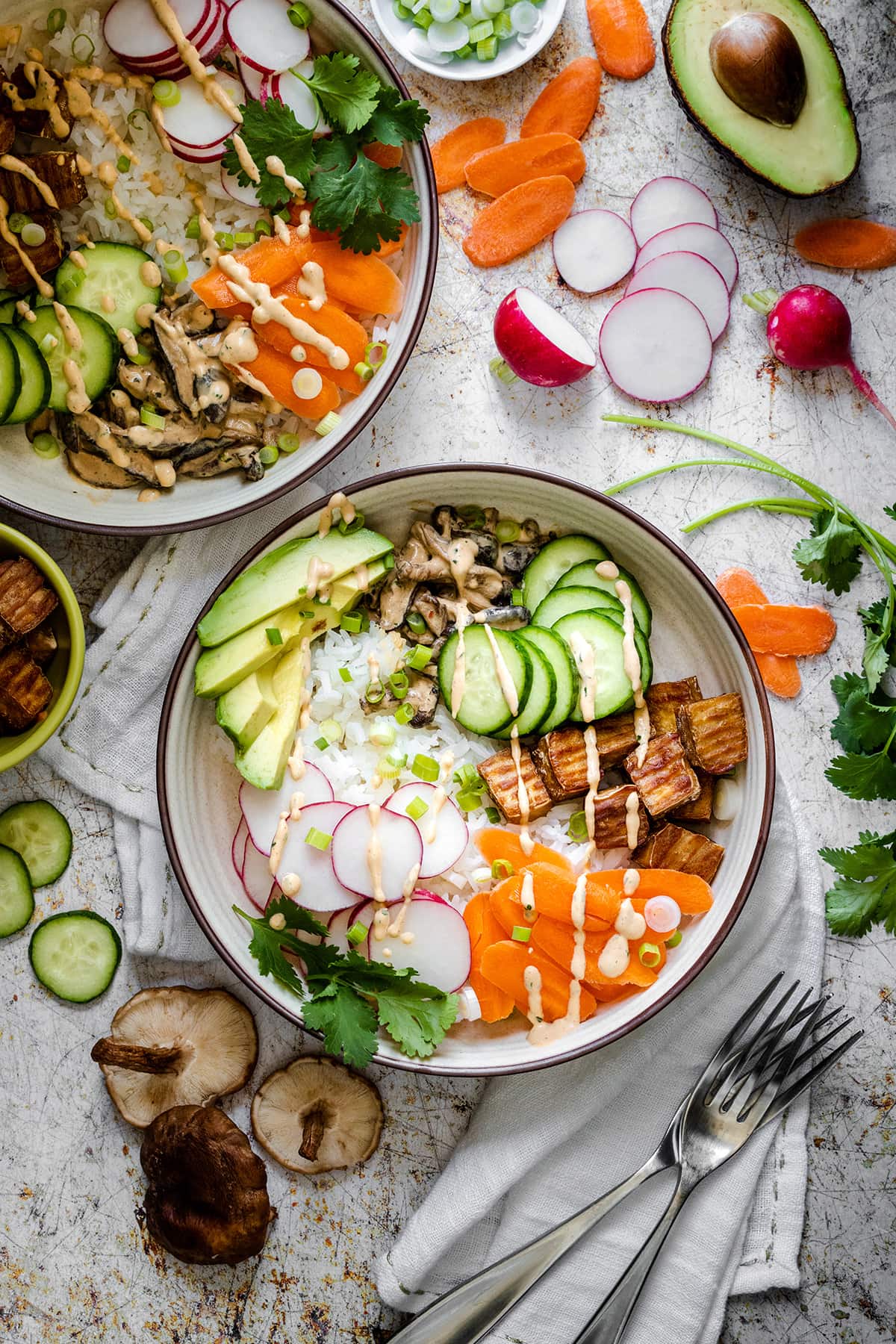 Two deconstructed sushi bowls with veggies and tofu surrounded by ingredients and two forks next to them