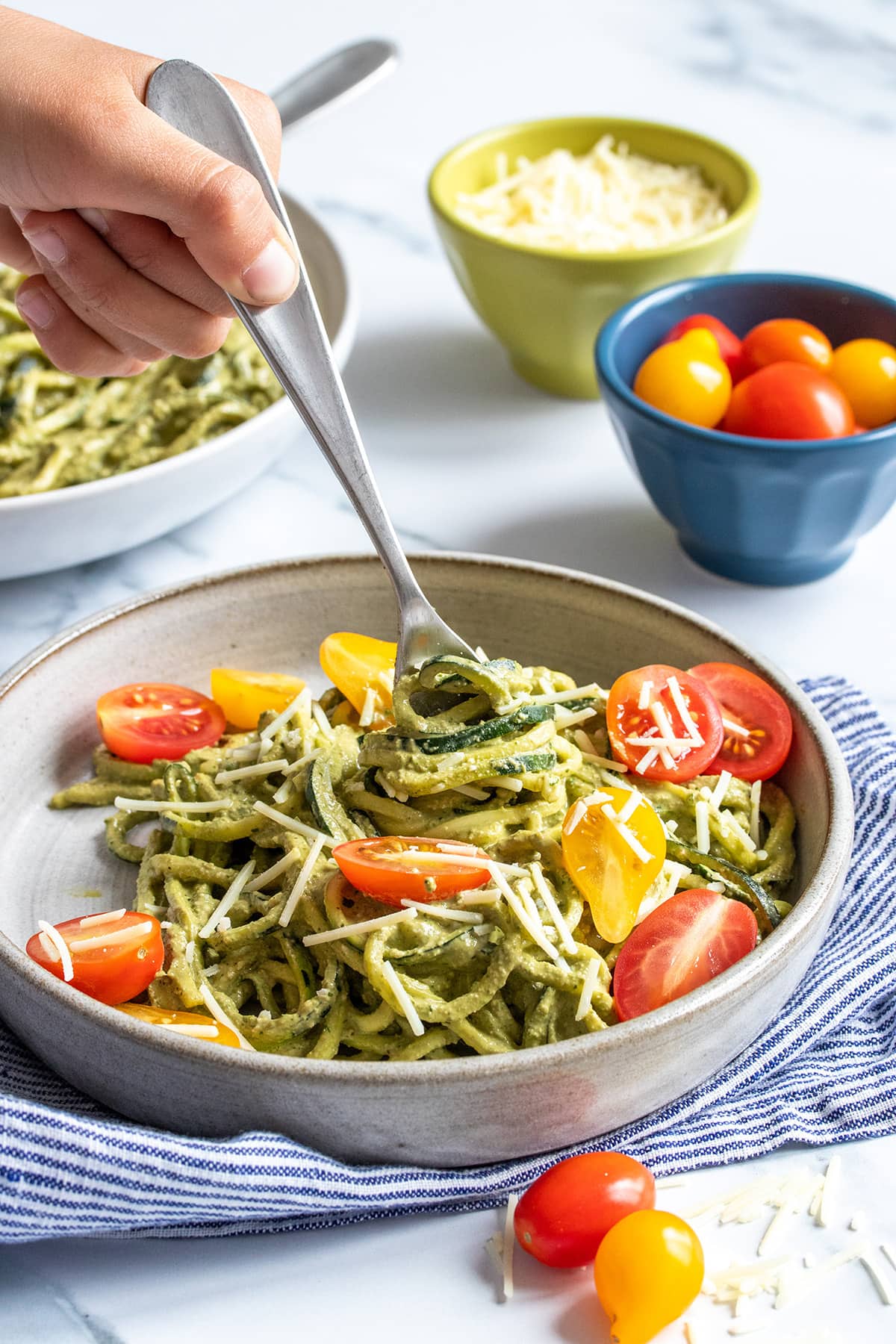 A hand holding a fork that is twisting pesto covered zucchini noodles onto it from a grey plate with tomatoes too.