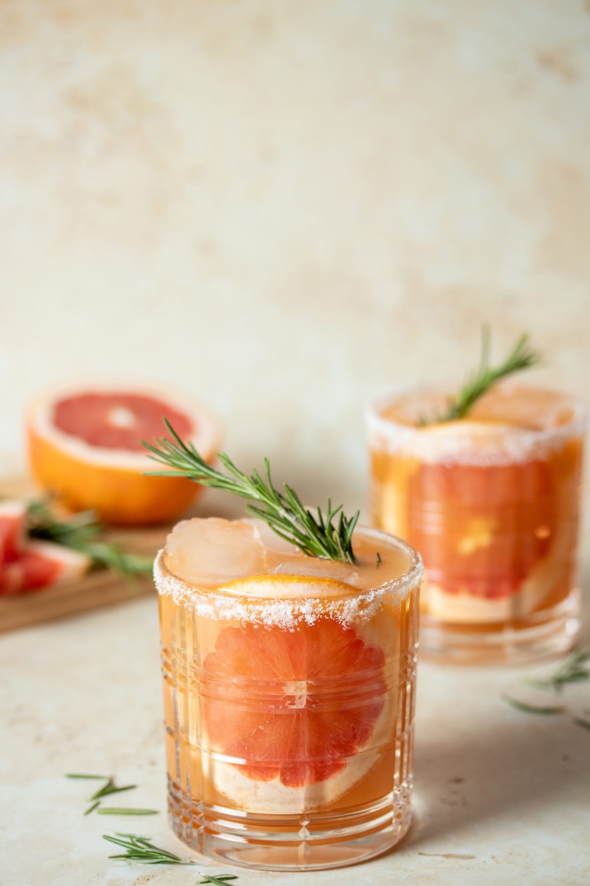 A cocktail glass with a round piece of grapefruit stuck to the front and a peach colors liquid with rosemary on top.