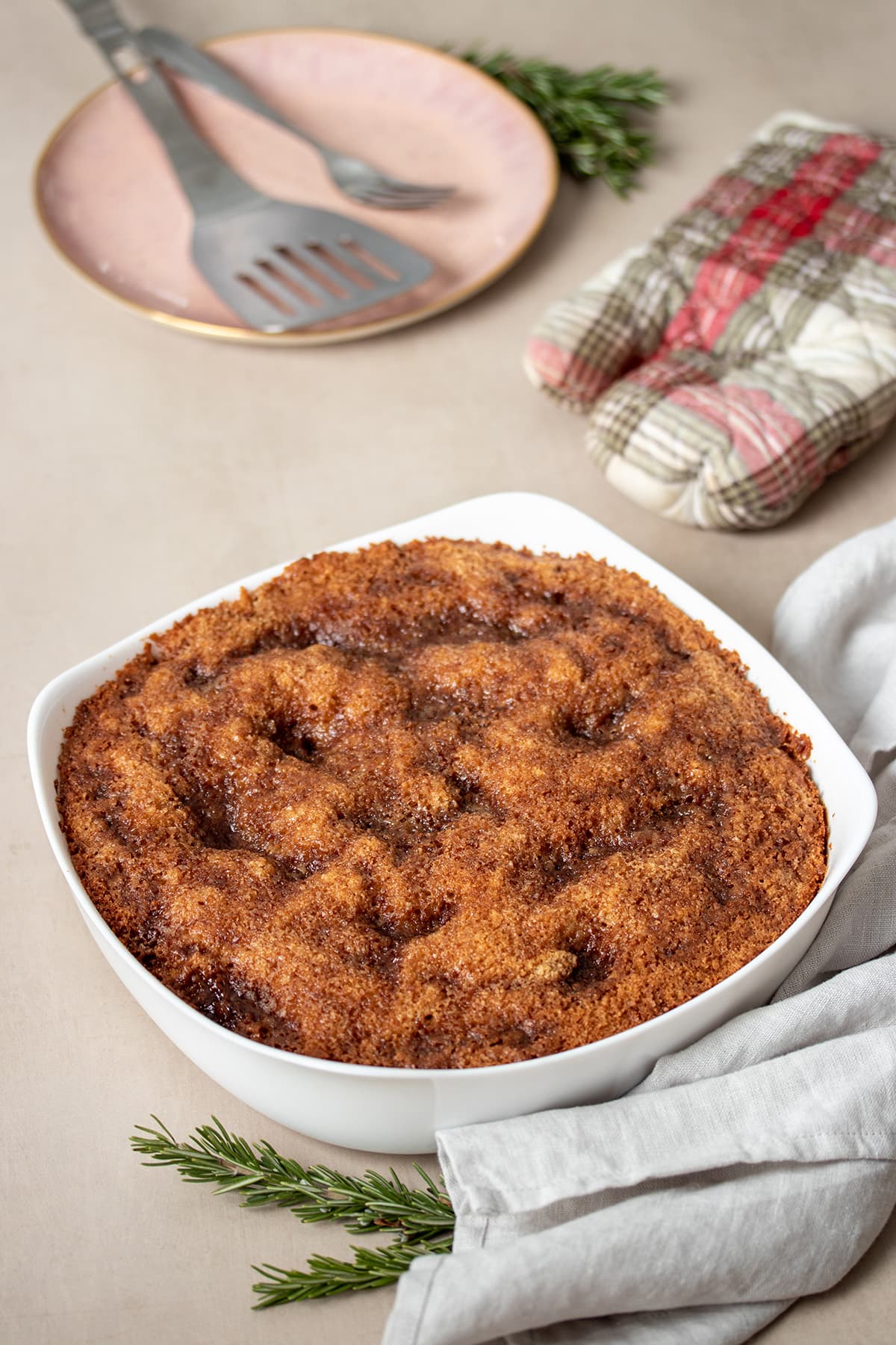 A white baking dish with baked coffee cake and a brown sugar streusel topping on a tan surface with a grey towel.