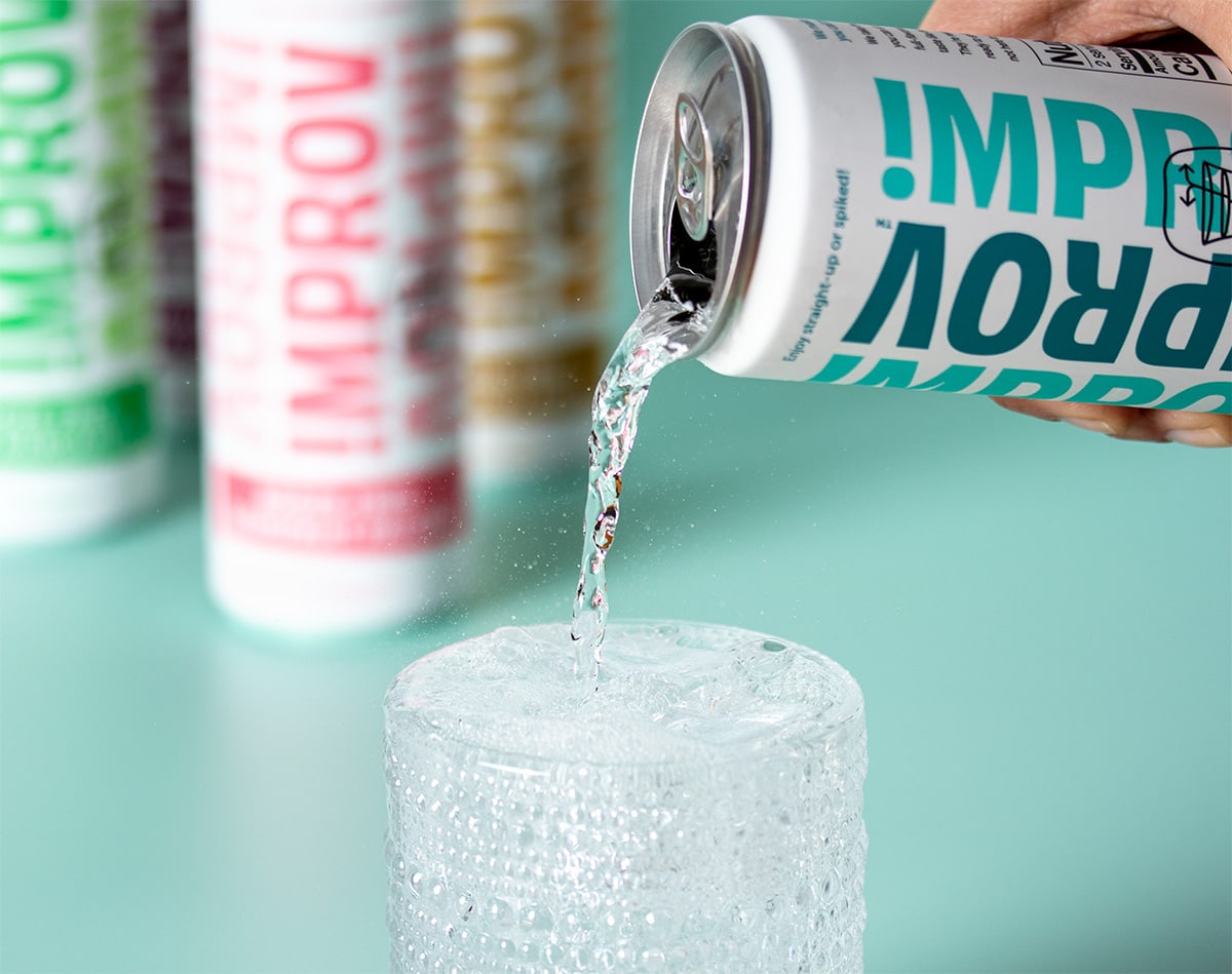 Hand pouring clear liquid from a white can with turquoise writing into a clear glass with texture on it on a mint green background.