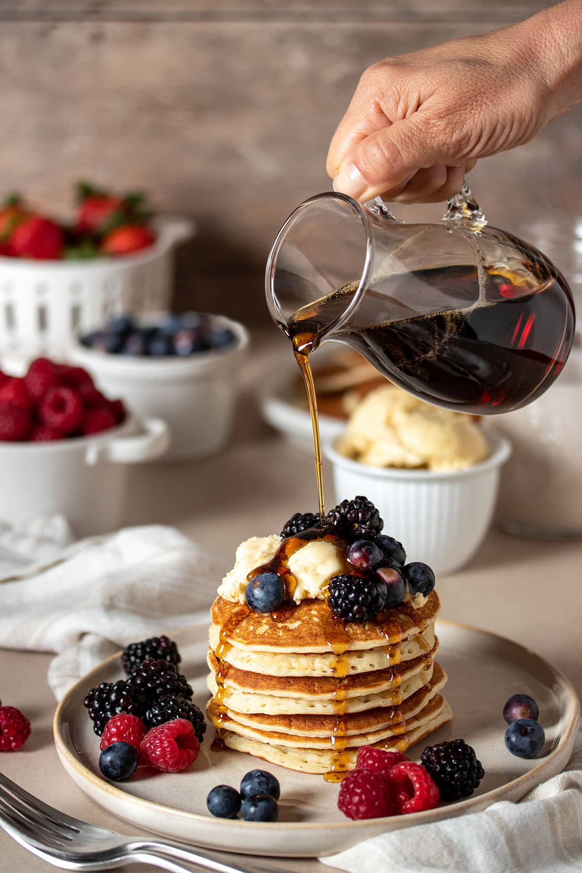 A hand pouring maple syrup from a glass container over a stack of pancakes with berries and butter.