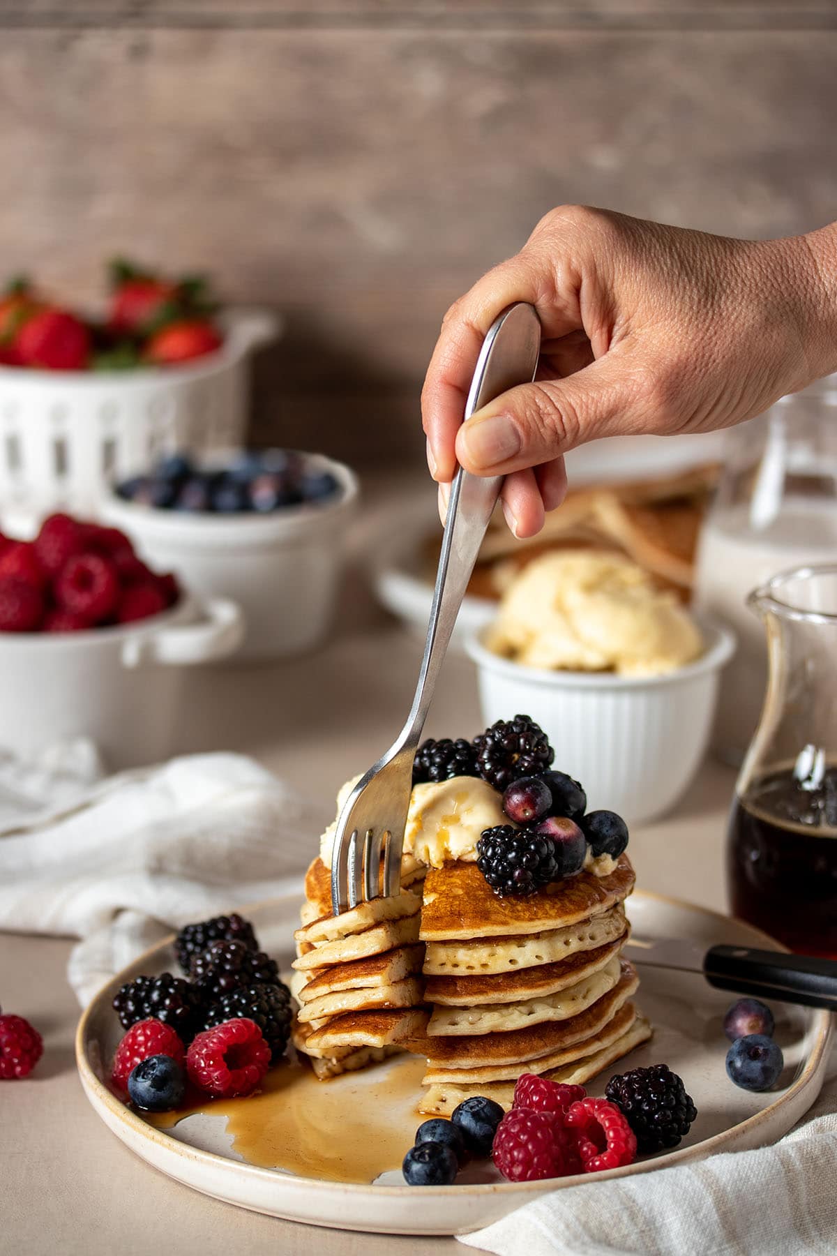 A hand holding a fork getting a bite out of a stack of pancakes topped with butter and berries.
