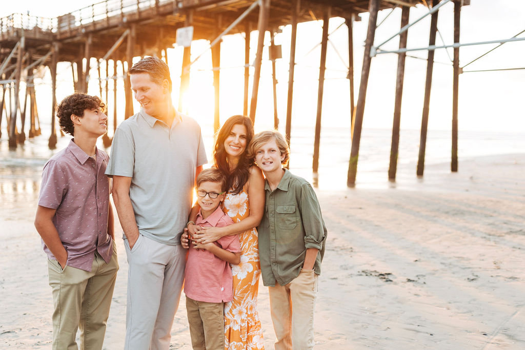 A family of 5 in front of a pier on the beach and the sun going down.
